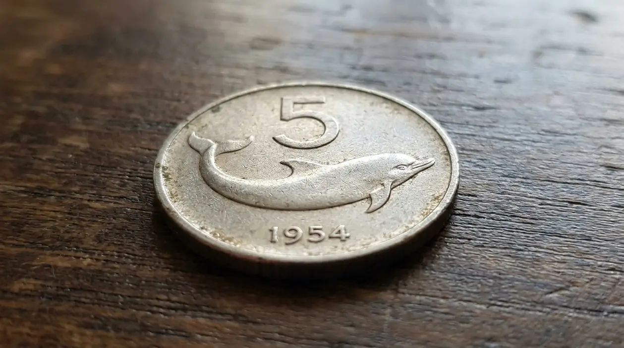 Moneta da 5 lire con delfino del 1954 appoggiata su un tavolo di legno
