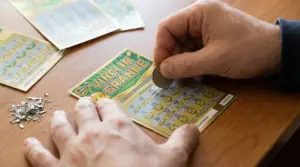 Mani che grattano un biglietto Gratta e Vinci su un tavolo, con altri tagliandi sullo sfondo.