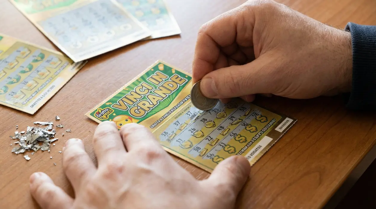 Mani che grattano un biglietto Gratta e Vinci su un tavolo, con altri tagliandi sullo sfondo.