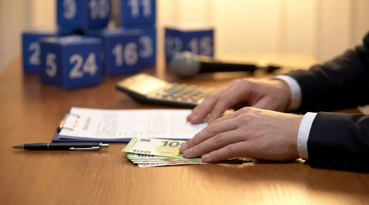 Mani su banconote in euro e documenti su un tavolo, con elementi che ricordano un quiz televisivo sullo sfondo