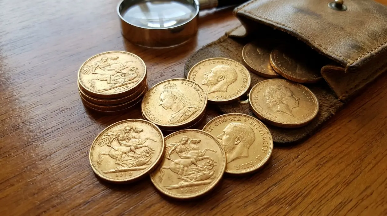 Sterline d’oro sparse su un tavolo accanto a una lente d’ingrandimento e a un portamonete in pelle