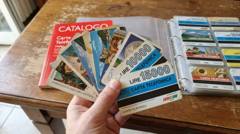 Mano che mostra una raccolta di vecchie carte telefoniche accanto a un catalogo e a un raccoglitore.