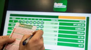 Persona compila una schedina del Superenalotto davanti a uno schermo con risultati e importi della vincita