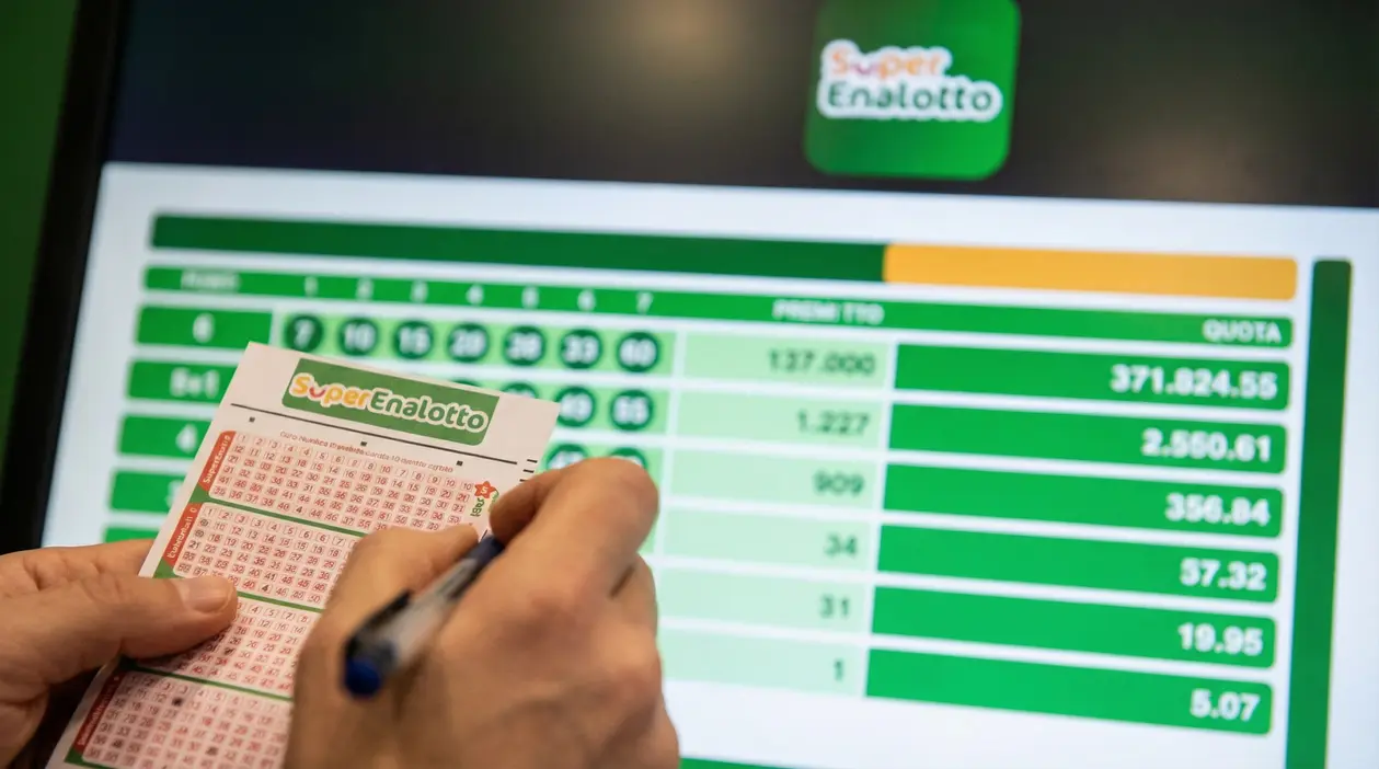 Persona compila una schedina del Superenalotto davanti a uno schermo con risultati e importi della vincita